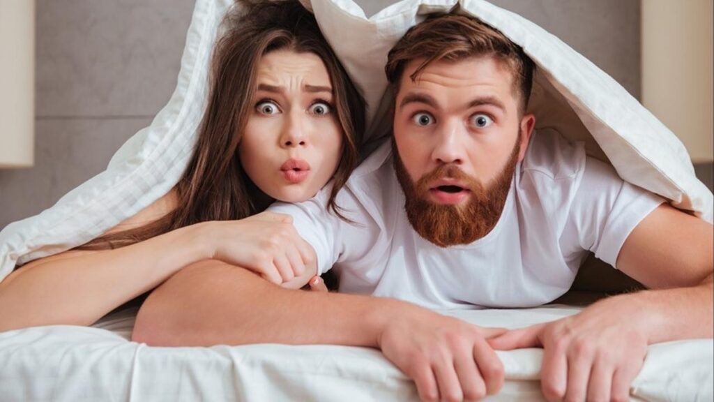 Man and Women lying on a bed under the covers looking out from the bottom of the bed with shocked faces