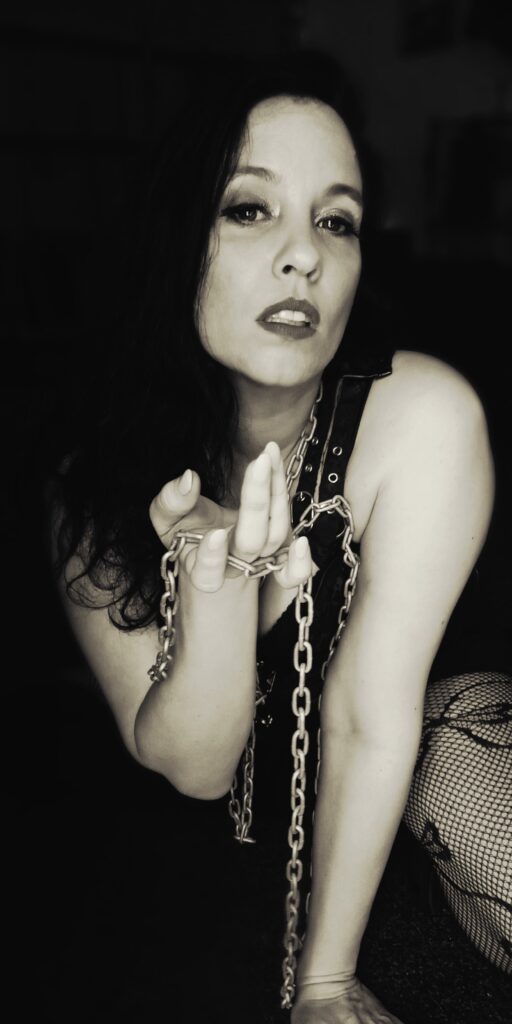 Mistress V holding a chain in her hand with a come hither motion implied